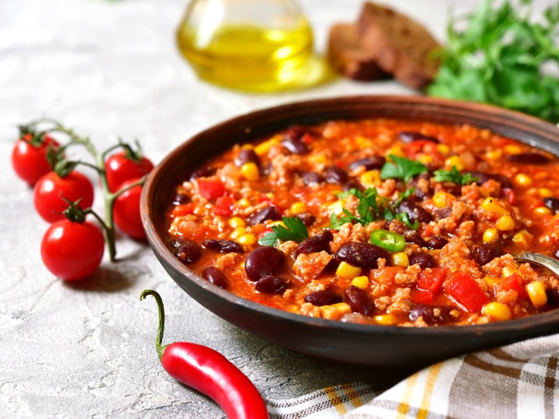 Ein tiefer Teller Chili con Carne mit Schokolade, darin ein Löffel, daneben Tomaten an der Rispe und eine Chilischote.