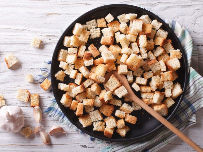 Croutons selber machen in der Pfanne, darin liegt ein Holzlöffel. Einige Croutons liegen als Deko außen um die Pfanne.