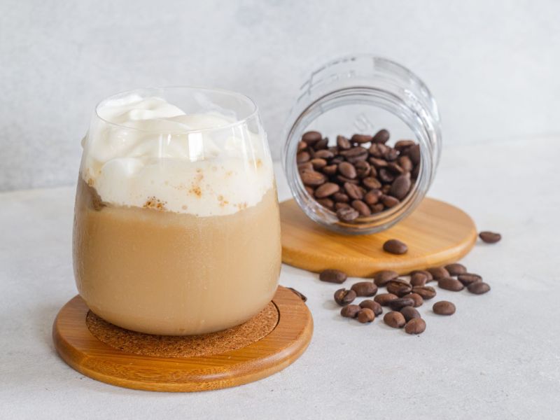 Ein Glas mit Espresso-Creme steht auf einem Untersetzer. Im Hintergrund ist ein Glas mit Kaffeebohnen umgekippt und liegt dekorativ im Bild.