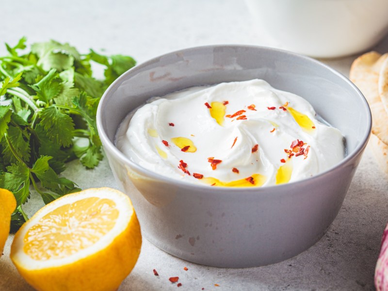 Eine Schüssel Feta-Knoblauch-Creme mit Öl und Chiliflocken garniert, daneben frische Kräuter und eine halbierte Zitrone sowie Fladenbrot.