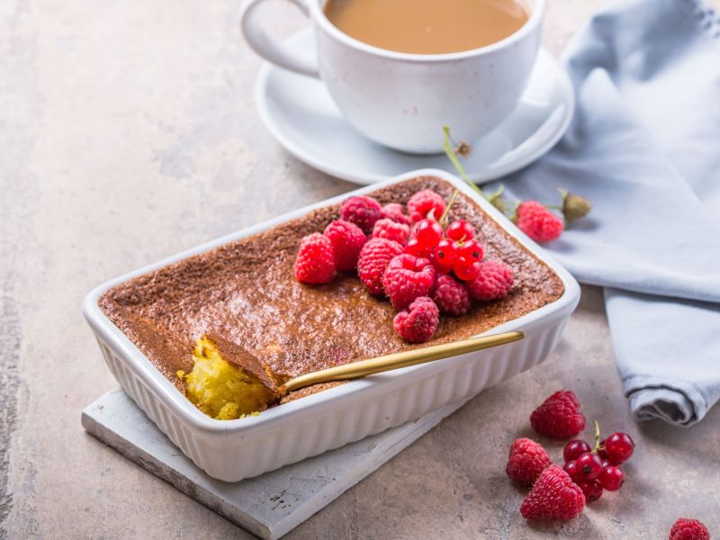 Gebackener Milchreis in einer Auflaufform, angestochen mit einem Löffel. Einige Himbeeren liegen als Dekoration auf dem Auflauf und daneben. Im Hintergrund steht eine Kaffeetasse.