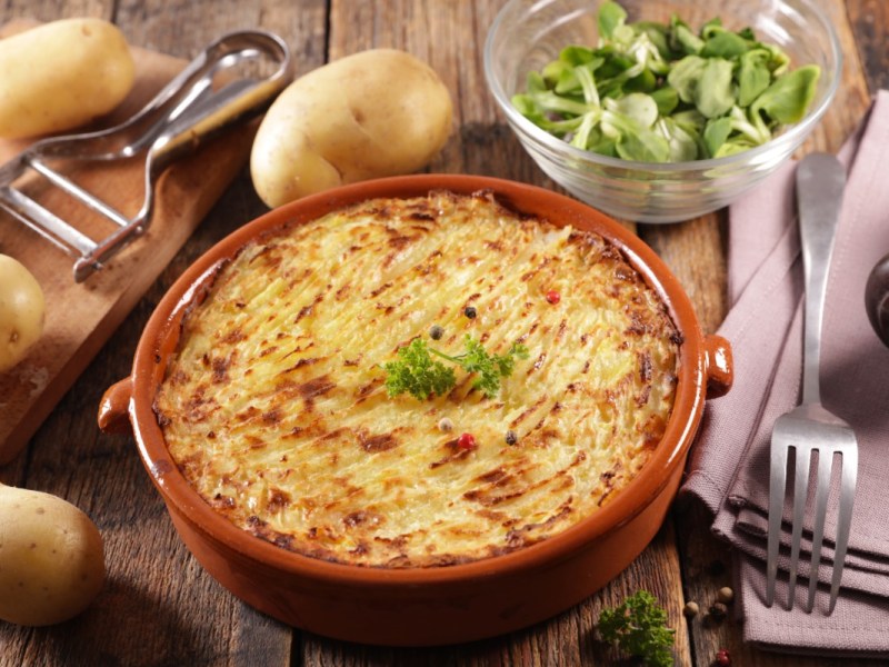 Gebackenes Kartoffelpüree in einer Schale. Drumherum liegen Kartoffeln, Küchengeräte und es steht eine Schüssel mit Feldasalat.