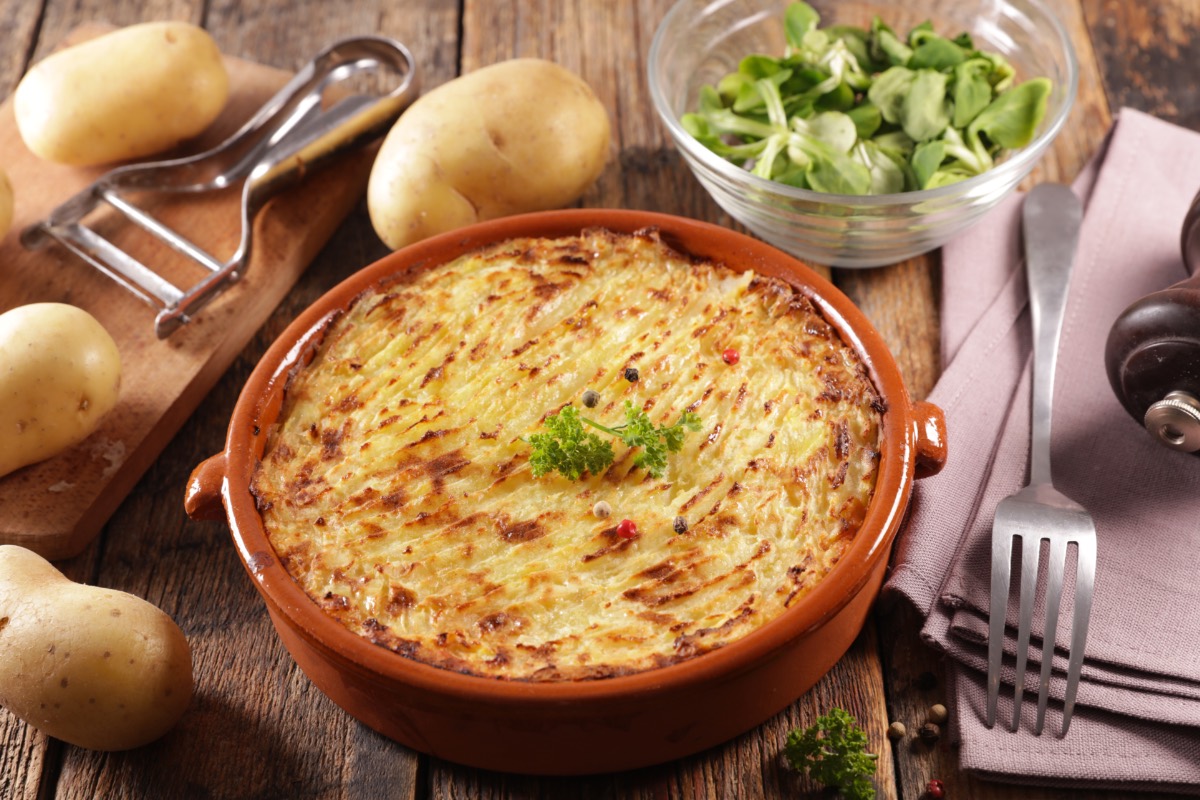 Gebackenes Kartoffelpüree in einer Schale. Drumherum liegen Kartoffeln, Küchengeräte und es steht eine Schüssel mit Feldasalat.