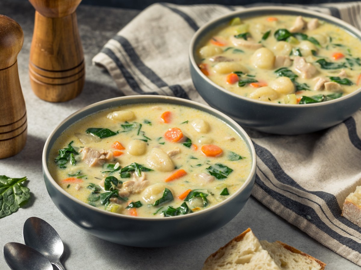 Zwei Schalen Hühnersuppe mit Gnocchi auf einem weißen Tisch. Eine von ihnen steht auf einem geschirrtuch. Daneben liegen brot, Löffel und zwei Pfeffermühlen.