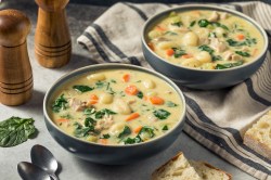 Zwei Schalen Hühnersuppe mit Gnocchi auf einem weißen Tisch. Eine von ihnen steht auf einem geschirrtuch. Daneben liegen brot, Löffel und zwei Pfeffermühlen.