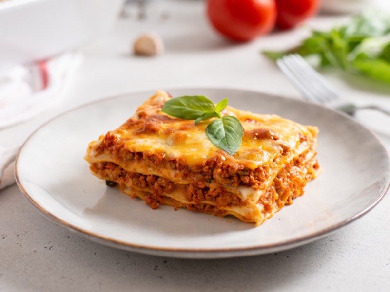 Ein Stück klassische Lasagne auf einem weißen Teller. Im Hintergrund sind frische Tomaten, Kräuter und die restliche Lasagne zu sehen.