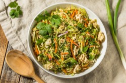 Knuspriger Ramen-Salat in einer weißen Schüssel, dekoriert mit etwas frischem Koriander. Daneben liegen ein Holzlöffel und eine Stange Frühlingszwiebel.