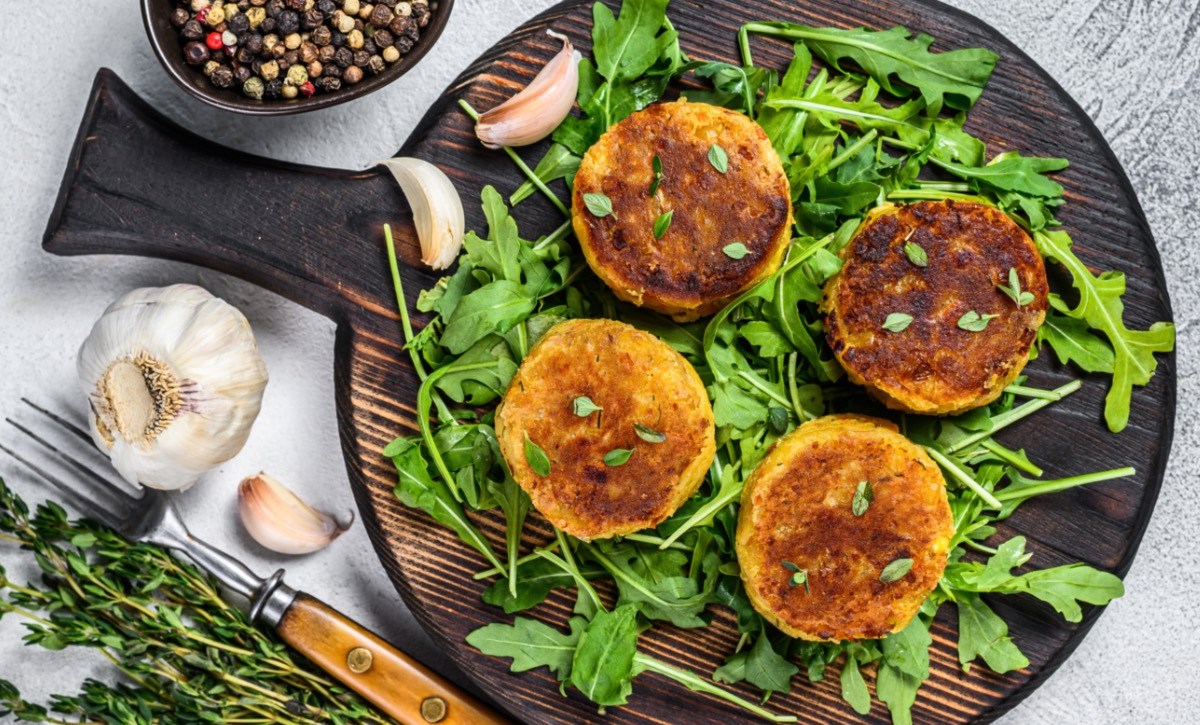 Vier Linsenfrikadellen auf einem Bett aus Rucola auf einem runden Holzbrett. Daneben steht eine Schale mit Pfeffer und es liegen Knoblauchzehen herum.