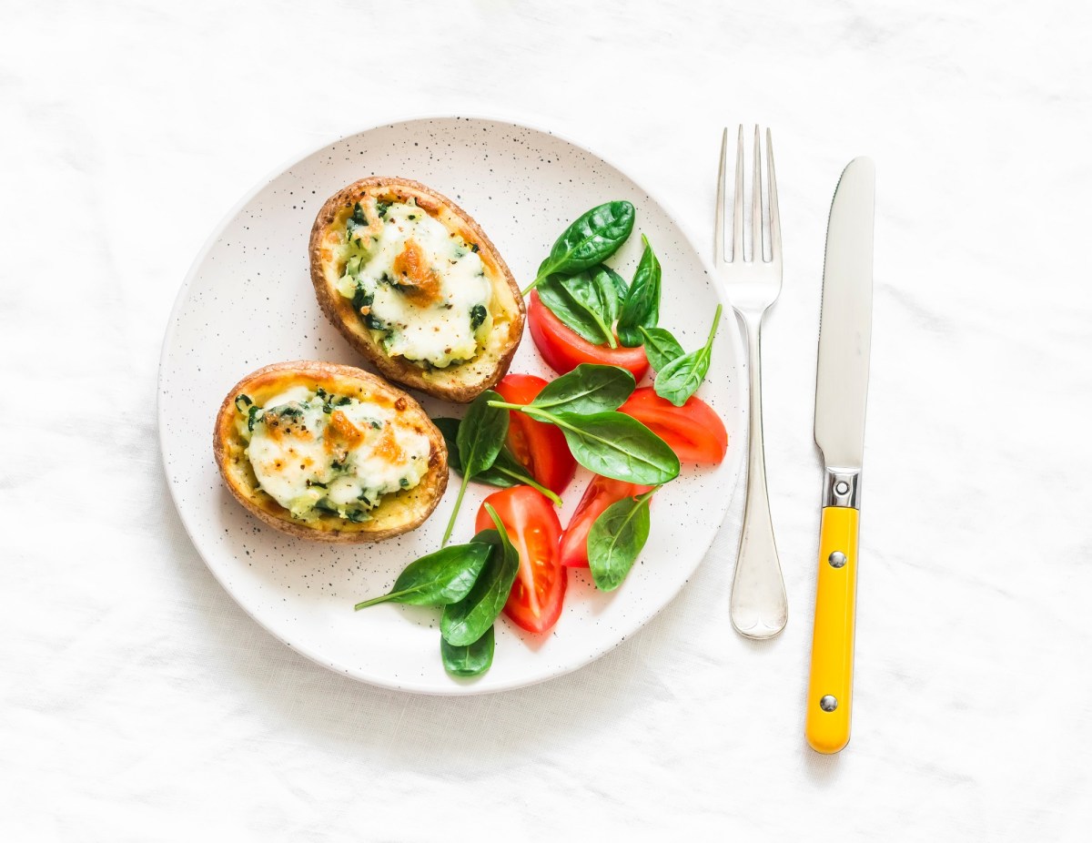 2 halbe Ofenkartoffeln mit Gorgonzola und Spinat auf einem Teller, auf dem auch ein Salat auf Feldsalat und Tomaten ist, daneben Messer und Gabel, heller Untergrund, Draufsicht.