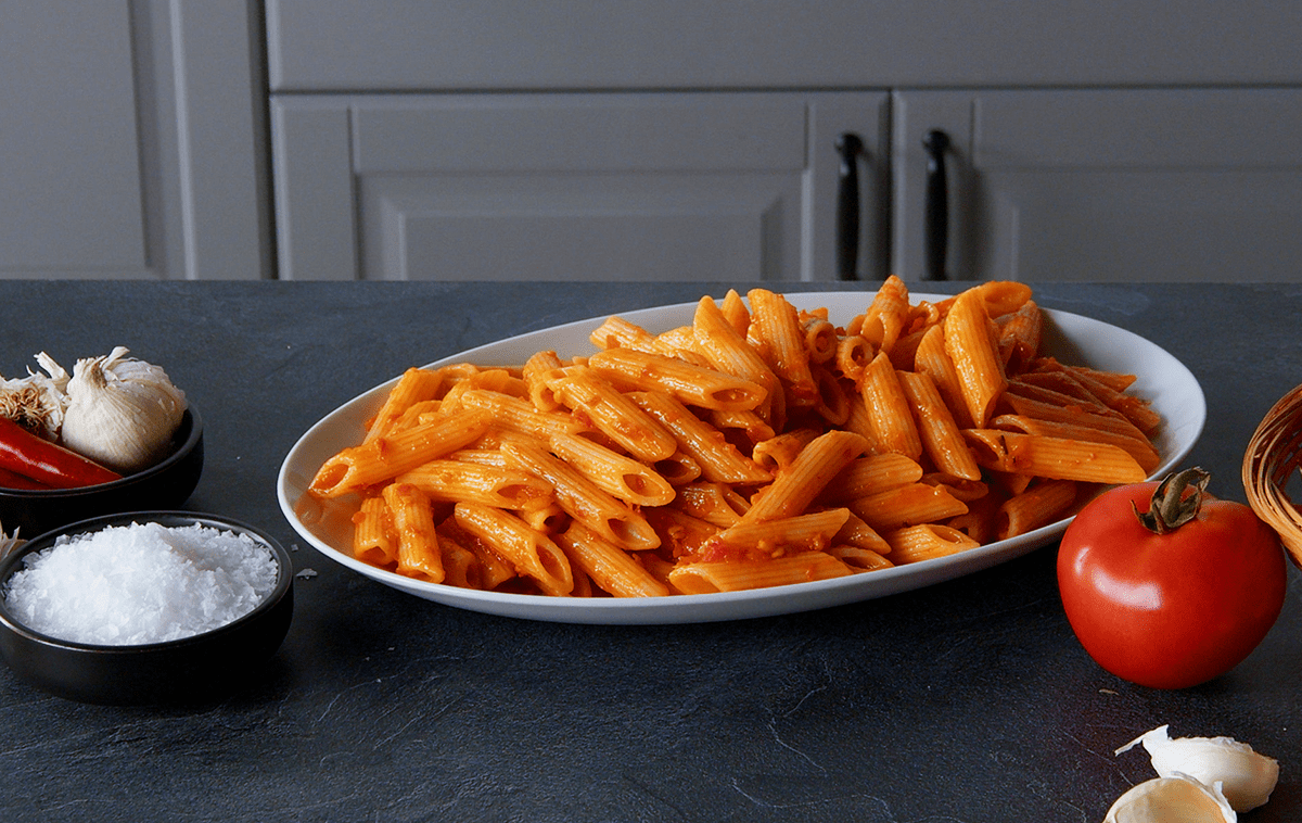 Penne Arrabiata, angerichtet auf einem Teller. Tomaten und Gewürze liegen außen herum.