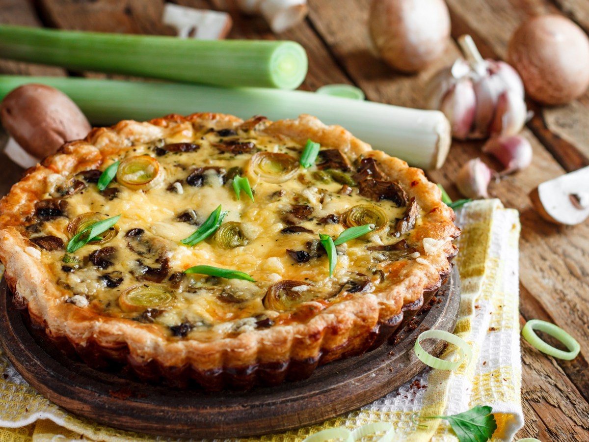 Eine Pilz-Lauch-Quiche, daneben ganze Pilze und Lauch auf Holzuntergrund.