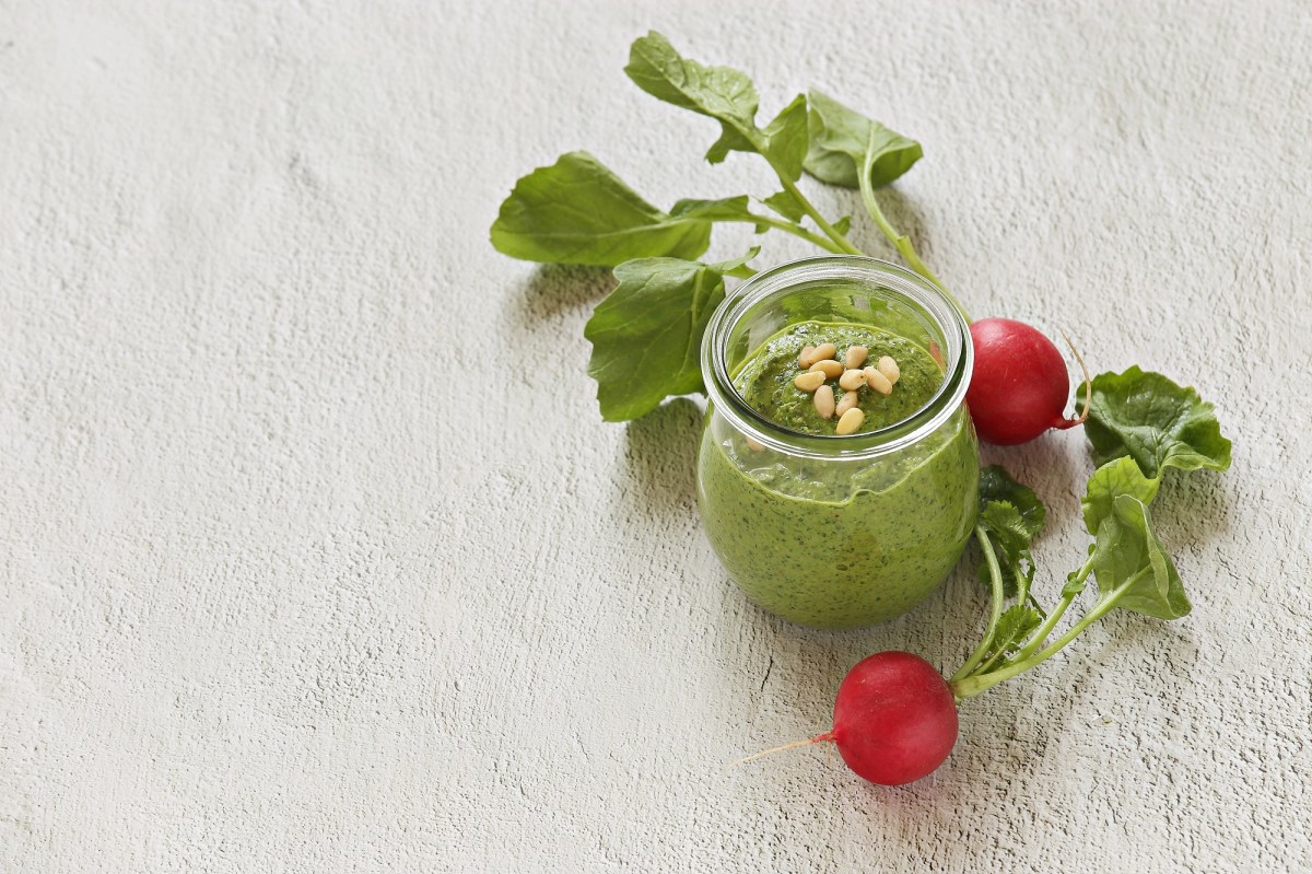 Ein Glas mit grünem Pesto und Radieschen daneben auf einem weißen Untergrund