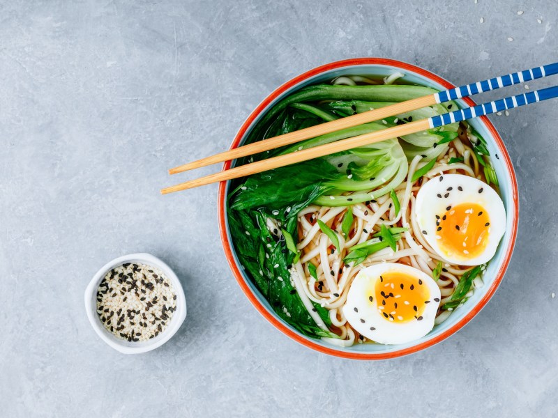 Eine Schüssel Erdnussbutter-Ramen mit halbiertem Ei und Pak Choi in der Draufsicht, darauf Stäbchen, daneben eine Schale Sesam.