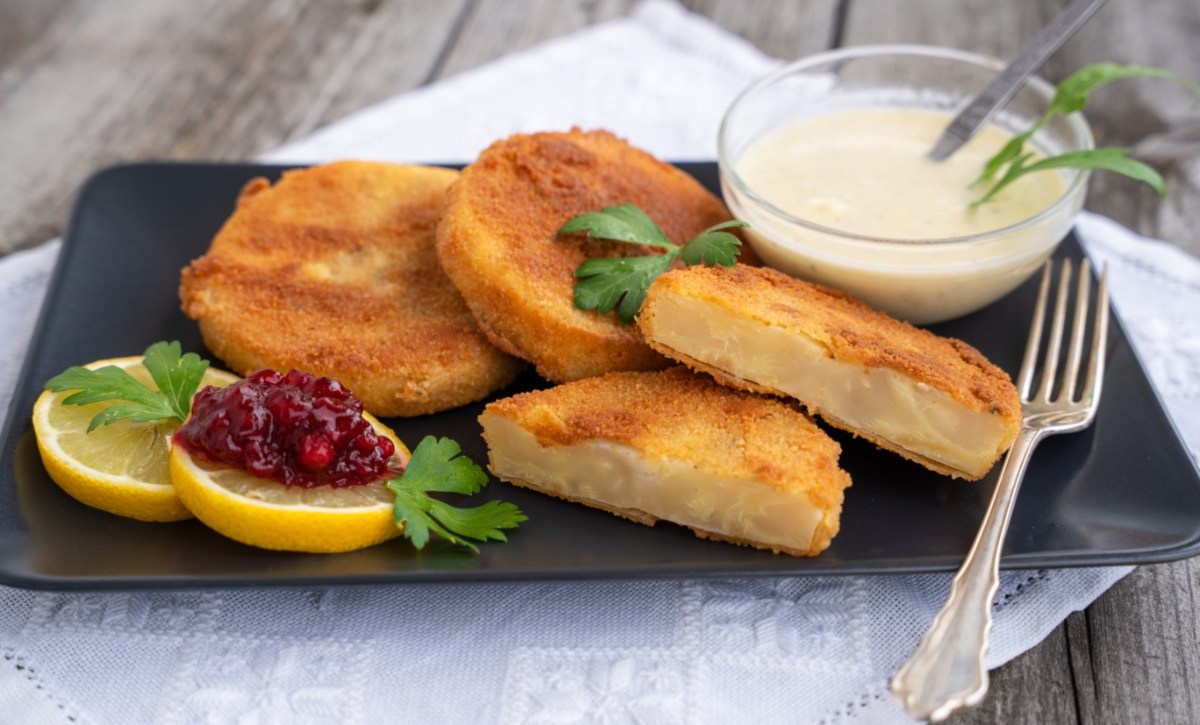 Mehrere Sellerieschnitzel auf einem schwarzen, rechteckigen Teller. Mit auf dem Teller sind zwei Zitronenscheiben, eine davon mit Preiselbeermarmelade, und ein Schälchen mit weißer Soße.