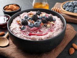 Süßer Couscous, serviert in einer dunklen Schale. Frische Heidelbeeren liegen als Deko obenauf. Schälchen mit Beeren, Honig und Mandeln stehen als Deko außen herum.