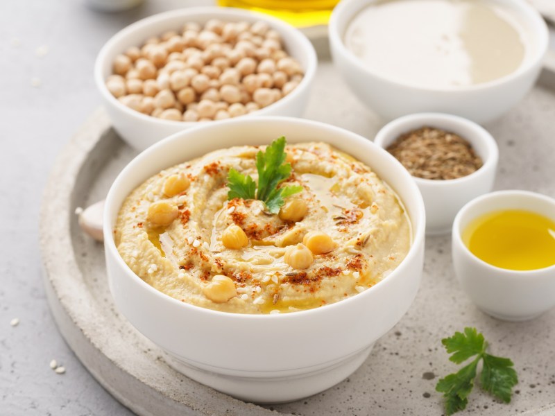 Tipps Hummus: eine weiße Schale Hummus mit Kichererbsen und Koriander sowie Sesam garniert, daneben je eine Schüssel rohe Kichererbsen, Öl, Gewürz und Kichererbsenwasser.