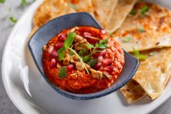 Eine Schale Tomate-Walnuss-Aufstrich, garniert mit Walnüssen, Kräutern und Granatapfelkernen, auf einem Teller, auf dem auch dreieckige Fladenbrote liegen, Nahaufnahme.