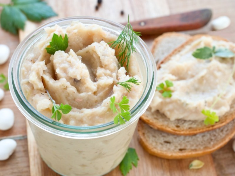 Weiße-Bohnen-Aufstrich im Glas. Daneben eine Scheibe Brot mit Aufstrich.