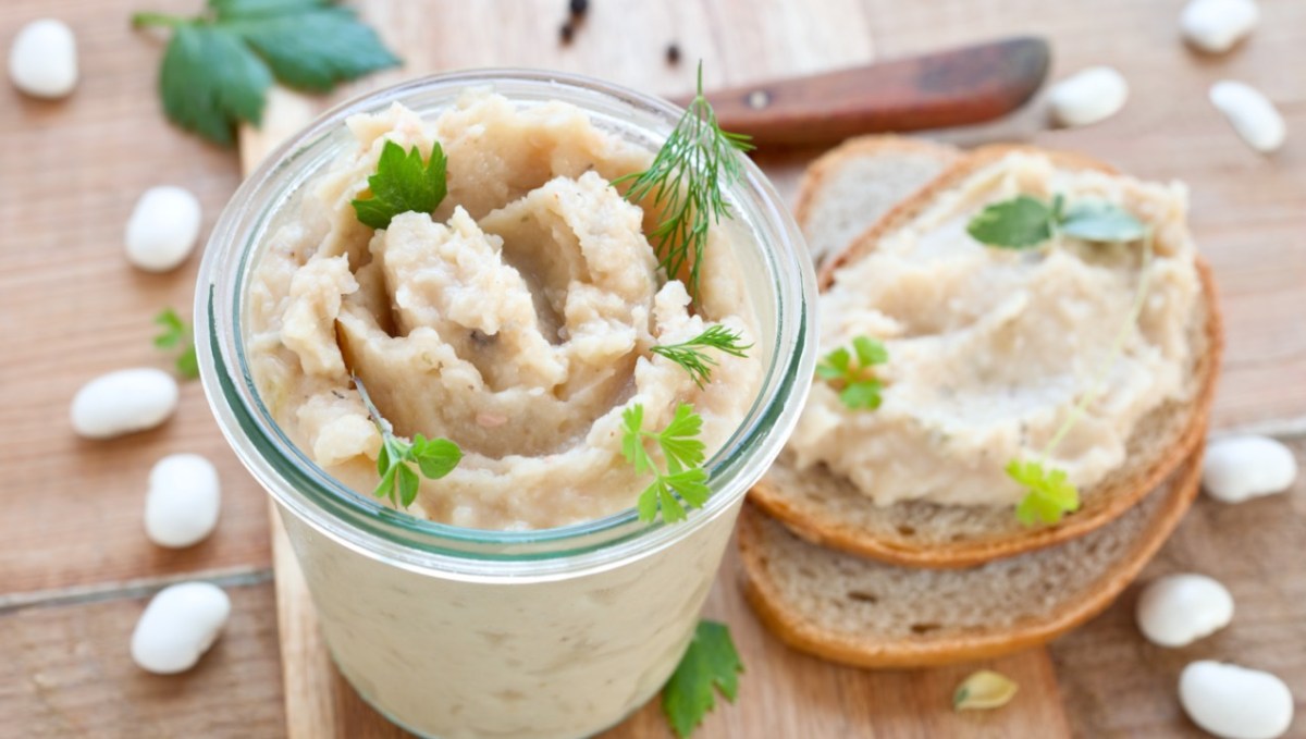 Weiße-Bohnen-Aufstrich im Glas. Daneben eine Scheibe Brot mit Aufstrich.