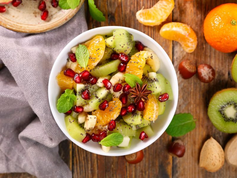 Winterlicher Obstsalat in einer Schüssel. Kiwi, Mandarinen und Orangen liegen als Deko außen herum.