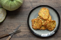 Ein Stapel Zucchini-Kartoffelpuffer auf einem Teller. Daneben liegen Besteck und Zucchini.