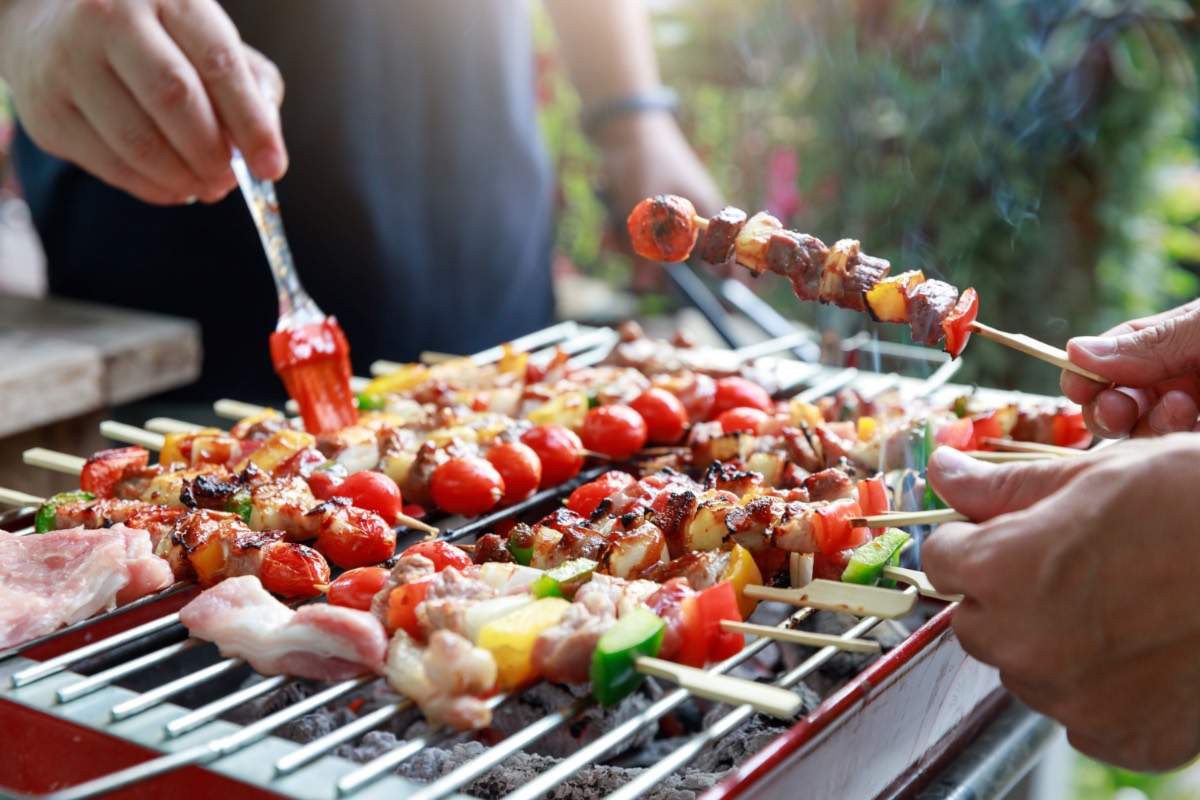 Angrillen: Grillgut auf einem Grill. Zwei Personen stehen um den Grill herum und halten Spieße und einen Pinsel in der Hand.