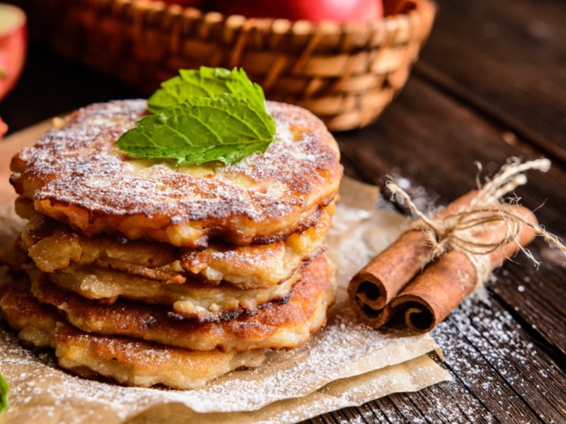 Ein Stapel Apfelpfannkuchen mit Puderzucker. Im Hintergrund sind frische Äpfel zu sehen, daneben liegen Zimtstangen.