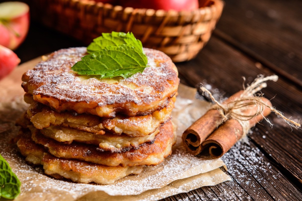 Ein Stapel Apfelpfannkuchen mit Puderzucker. Im Hintergrund sind frische Äpfel zu sehen, daneben liegen Zimtstangen.