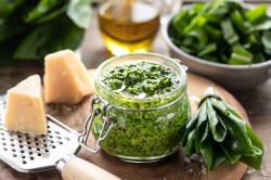 ein Glas Bärlauchpesto, daneben frischer Bärlauch und Parmesan sowie eine Käsereibe und eine Olflasche.