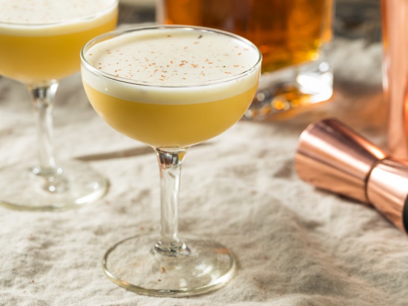 Zwei Gläser Bourbon Flip Cocktail mit Cocktailshaker auf hellem Tischtuch.