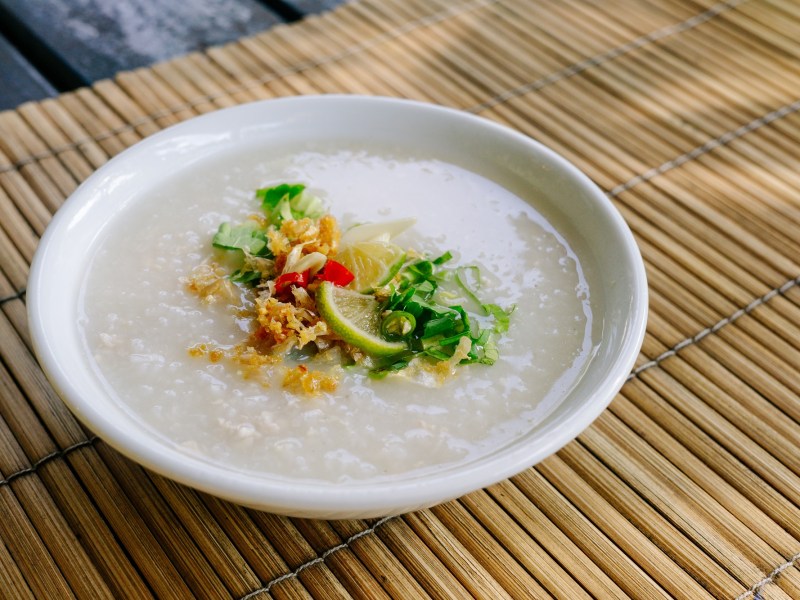 Congee in einer weißen Schale auf einer Bambusmatte. Als Toppings sind Röstzwiebeln, Frühlingszwiebeln und Limetten eingesetzt.