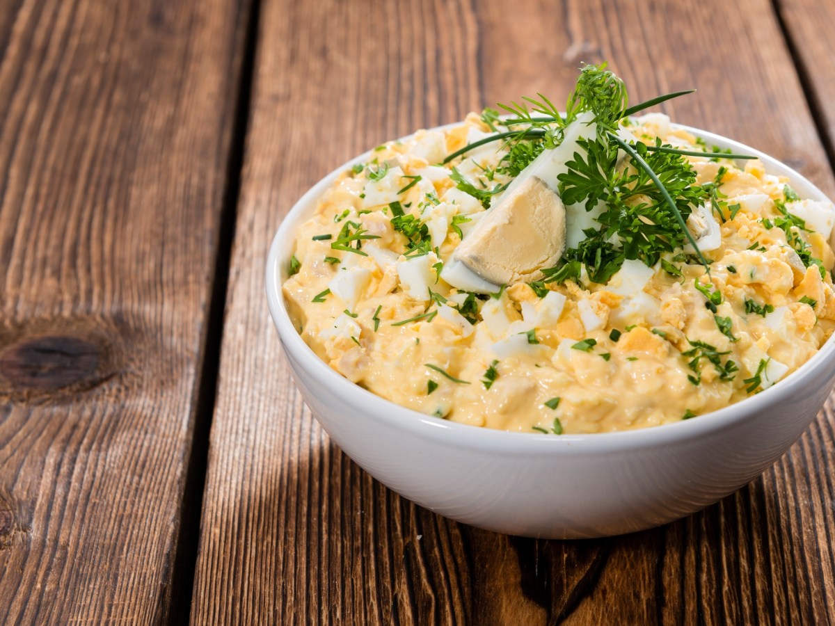 Eiersalat in einer weißen Schale, die auf einem dunklen Holztisch steht. Garniert ist er mit einem Stück Ei und frischen Kräutern.