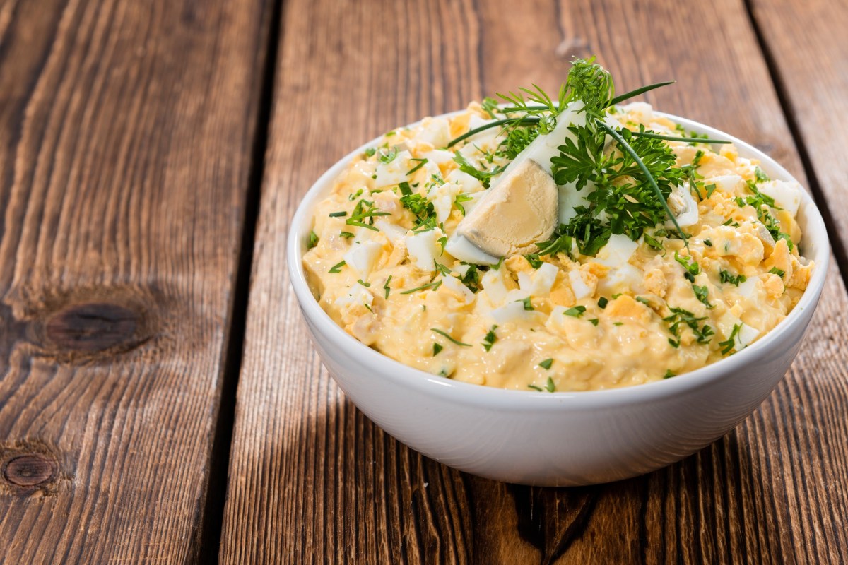 Eiersalat in einer weißen Schale, die auf einem dunklen Holztisch steht. Garniert ist er mit einem Stück Ei und frischen Kräutern.