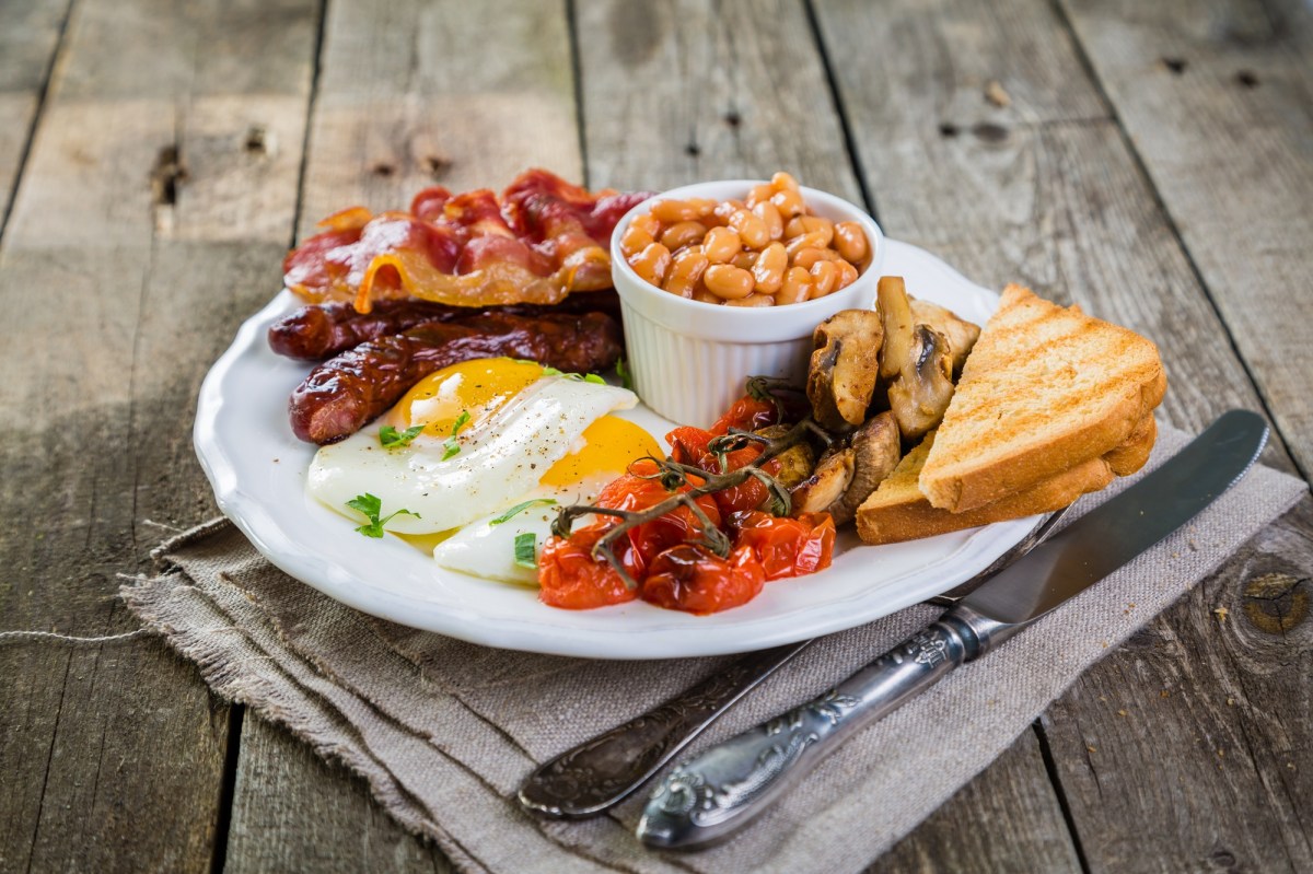 English Breakfast mit Bacon, Würtchen, Spiegeleiern, Baked Beans, Tomaten, Pilzen ud Toast auf einem weißen Teller. Daneben Messer und Gabel.