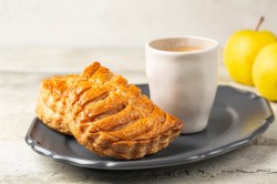 Französische Apfeltaschen auf einem schwarzen Teller. Daneben eine weiße Tasse. Im Hintergrund zwei gelbe Äpfel.