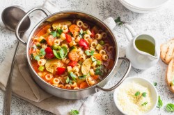 Ein Topf mit Frühlings-Minestrone in der Draufsicht. Daneben liegt eine Kelle und es stehen ein Schälchen mit Käse und eine Kanne mit Öl herum.