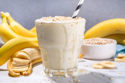 Ein Glas Frühstücksshake mit Bananen und Haferflocken. Im Hintergrund sind Bananen uds eine Schale mit Haferflocke zu sehen.