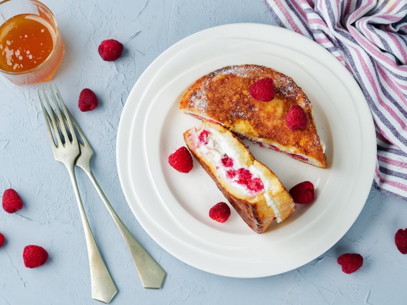 Gefüllte Arme Ritter mit Frischkäse und Marmelade und Himbeeren auf einem Teller, drumherum Messer und Gabel und frische Himbeeren, Draufsicht.