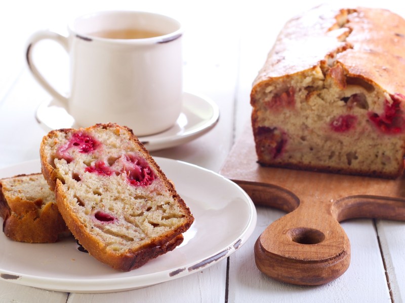 Ein angeschnittener Himbeerkuchen mit weißer Schokolade auf einem Brett, daneben ein Teller mit zwei Stücken Kuchen und eine Tasse.