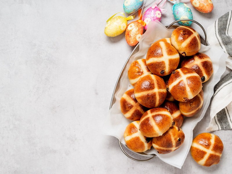 Hot Cross Buns in einer Metallschüssel, daneben bunte Ostereier und ein Geschirrtuch.