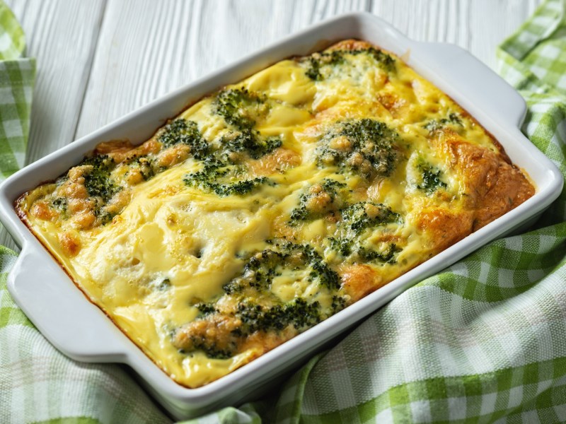 Kartoffel-Brokkoli-Gratin ohne Vorkochen in einer Auflaufform, darum ist ein grün-weiß kariertes Geschirrtuch drapiert.