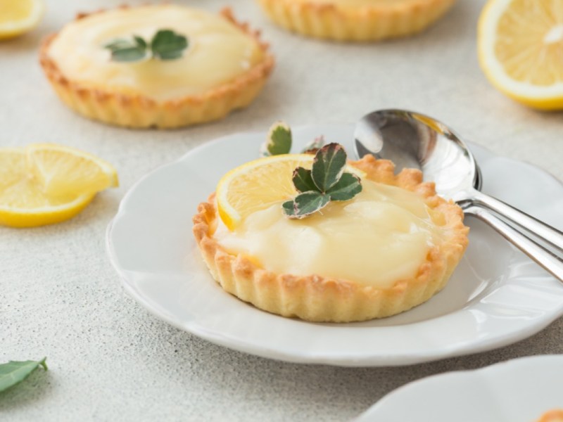 Zwei Lemon Curd Tartelettes auf Tellern. Neben einem liegt Besteck, im Hintergrund aufgeschnittene Zitronen.