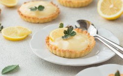 Zwei Lemon Curd Tartelettes auf Tellern. Neben einem liegt Besteck, im Hintergrund aufgeschnittene Zitronen.