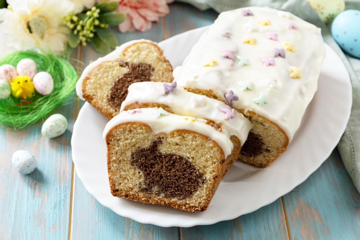 Ein angeschnittener Osterhasen-Kuchen auf einem Teller. Im Hintergrund liegen Blumen und bunte Ostereier. Ein Küken sitzt in einem Nest mit Eiern daneben.