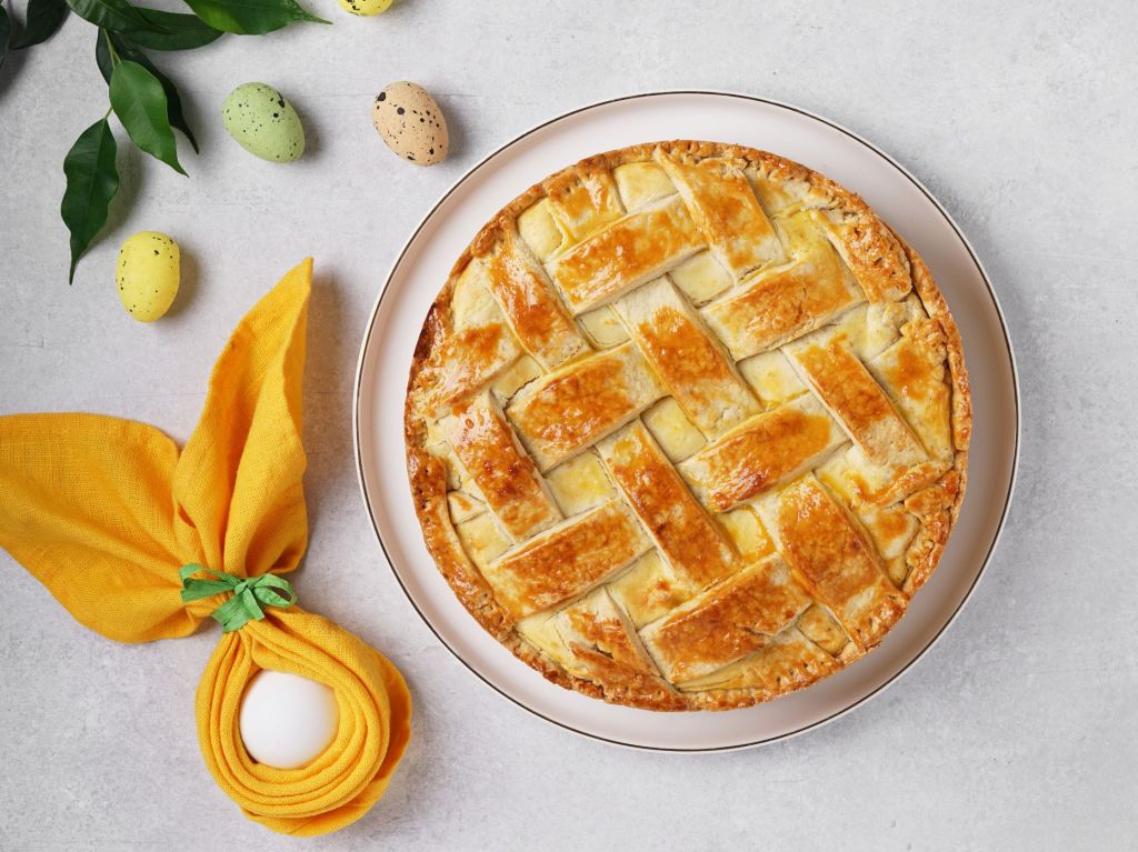 Pastiera napoletana: italienischer Osterkuchen in Gitteroptik auf einem Teller. Daneben liegt ein Osterei mit Serviettenohren als Dekoration.