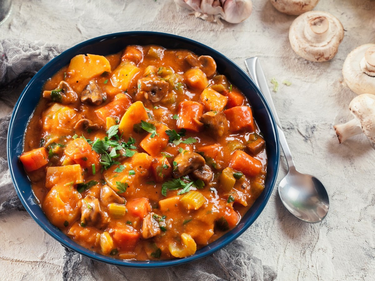 Ein Teller Pilzgulasch mit Möhren und Sellerie, daneben ein Löffel und Pilze, Draufsicht.