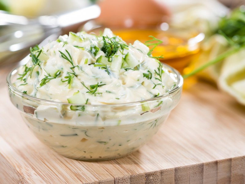 Remoulade in einer Glasschale, die auf einem Holzbrett steht. Dahinter sind unscharf die Zutaten zu erkennen.