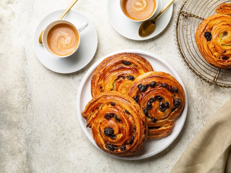 Rosinenschnecken auf einem weißen Teller. Dieser steht auf einem beigen Tischtuch. Daneben eine Tasse Kaffee und ein Gitter mit noch mehr Rosinenschnecken.