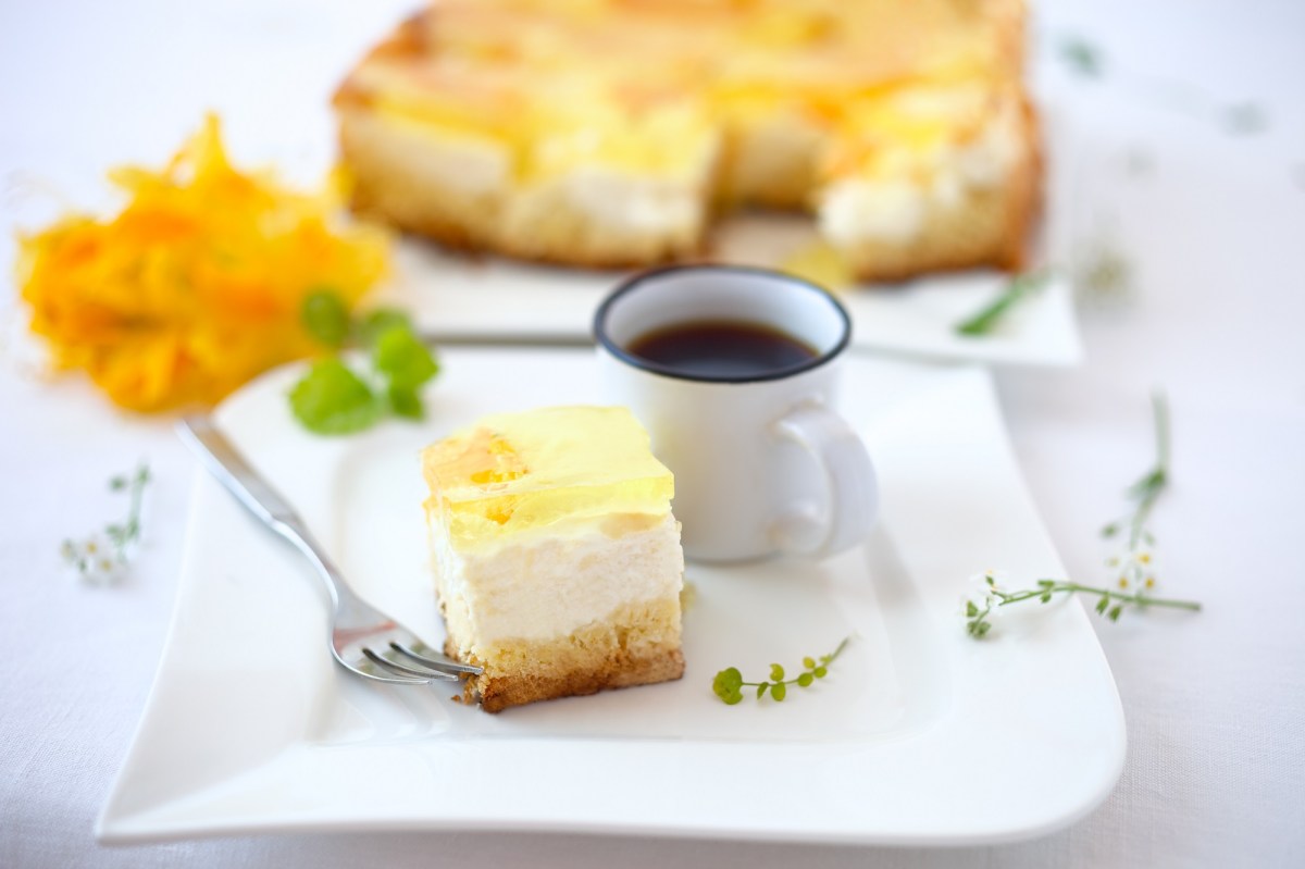 Ein Stück Schmankuchen vom Blech mit Früchten auf einem Teller, daneben eine Kaffeetasse, dahinter der Kuchen.