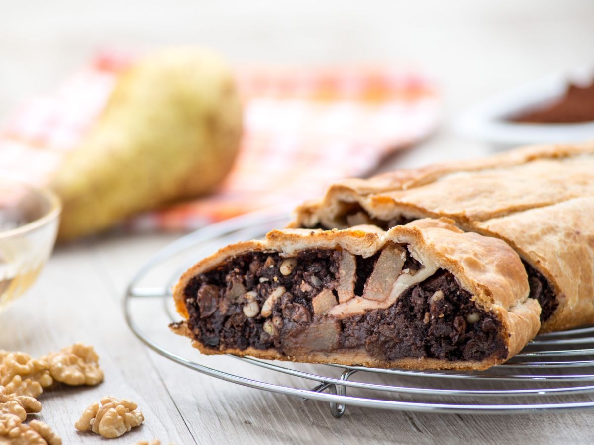 Schoko-Birnen-Strudel, in Scheiben geschnitten auf einem Gitter liegend. Im Hintergrund und daneben liegen eine Birne und Walnüsse als Deko.